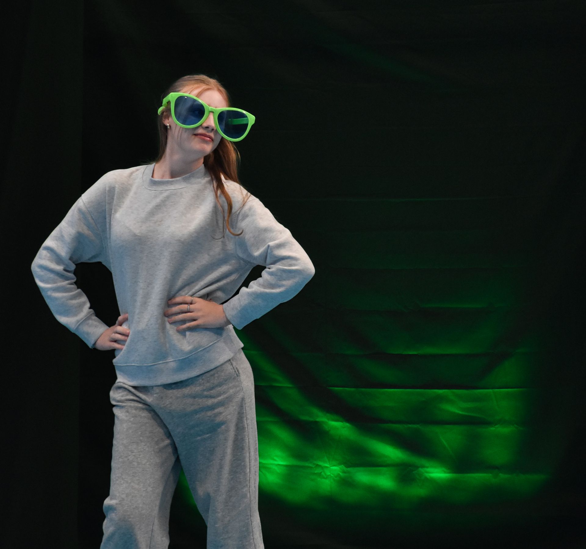 A teenage girl in oversized green sunglasses and gray sweats poses against a dark backdrop with green light.