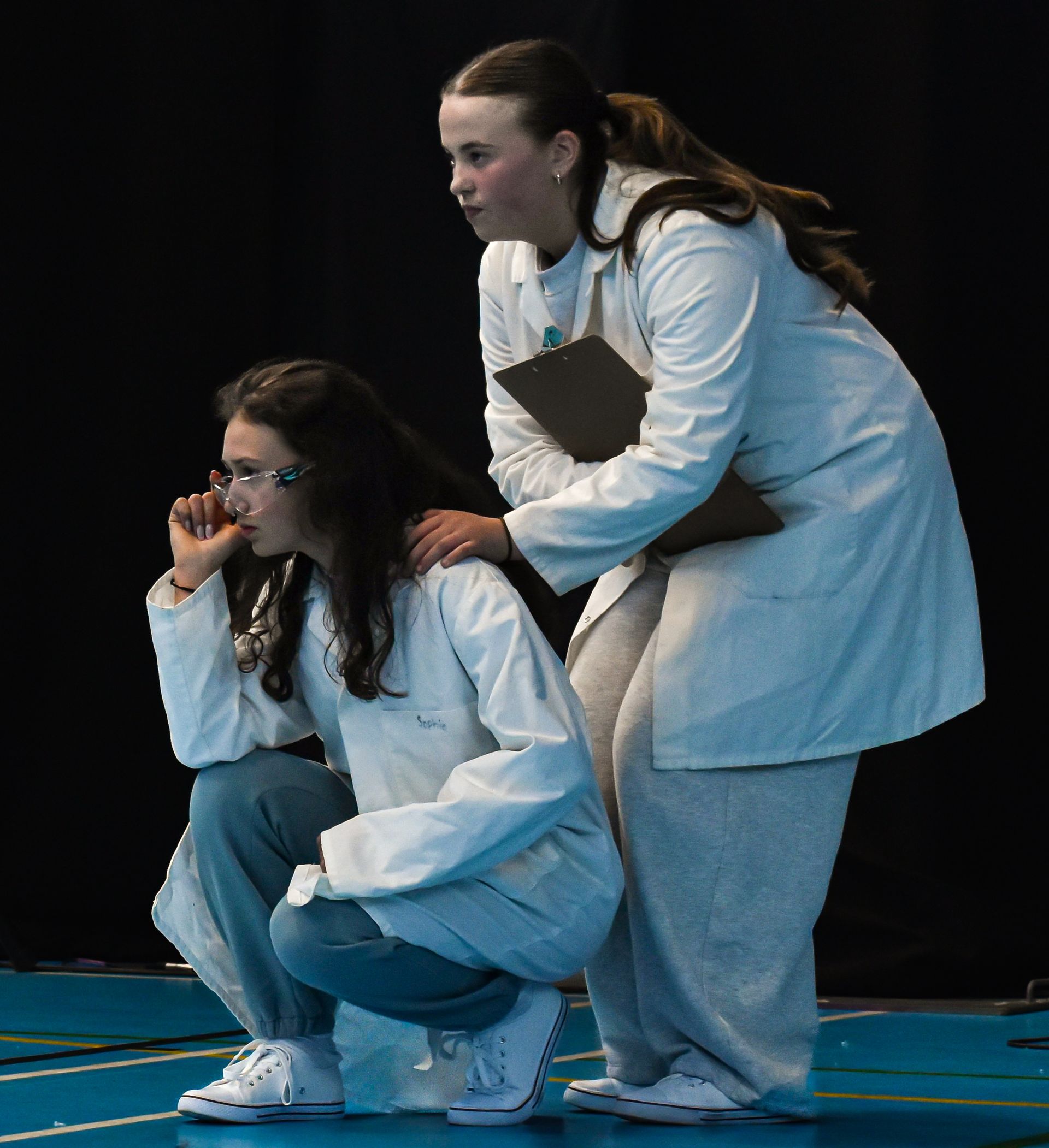 Two teenagers in white lab coats crouch on a blue stage, one supporting the other while both face left.