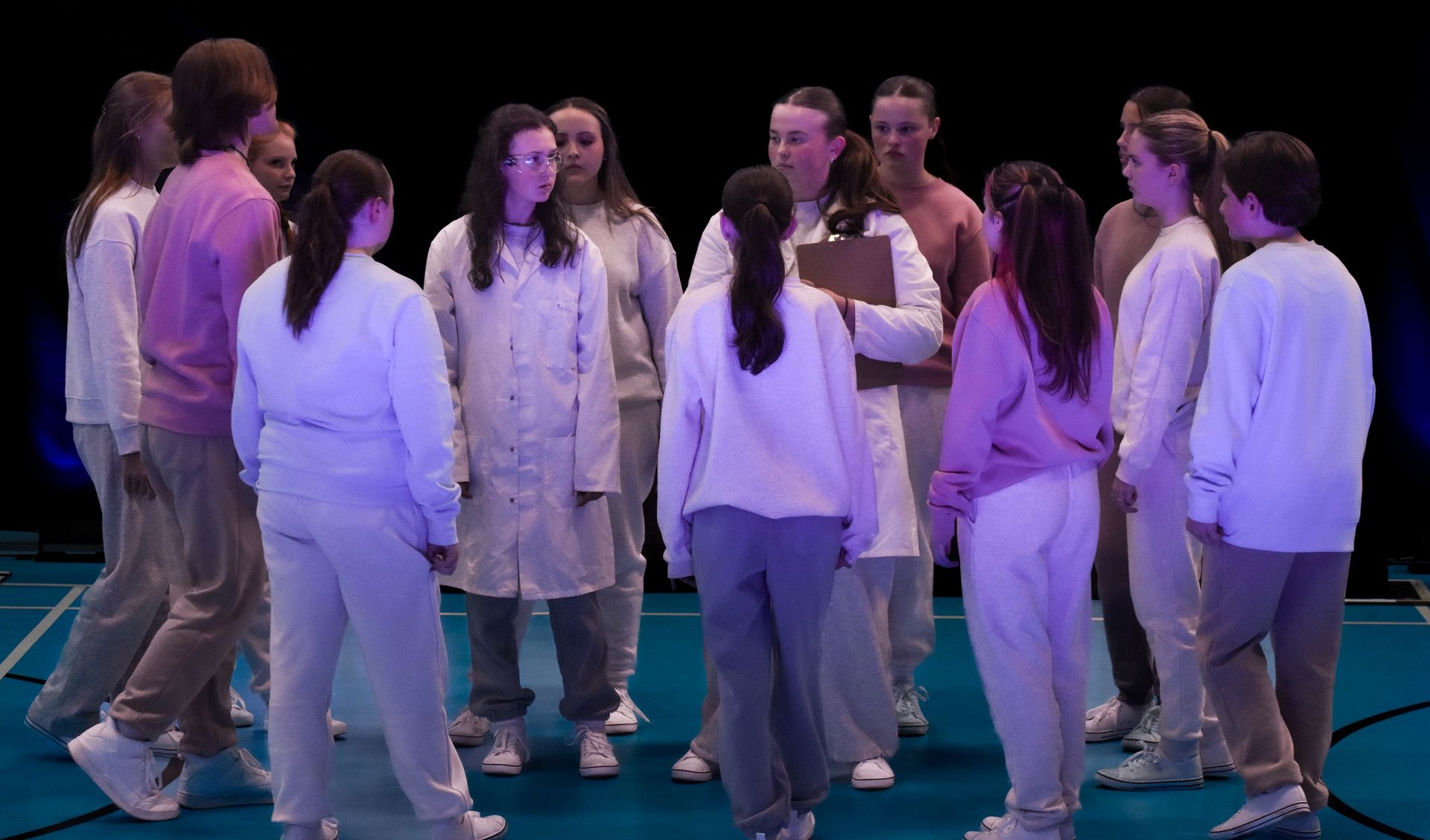 Group of people in white and light-purple outfits standing in a circle on a blue stage.