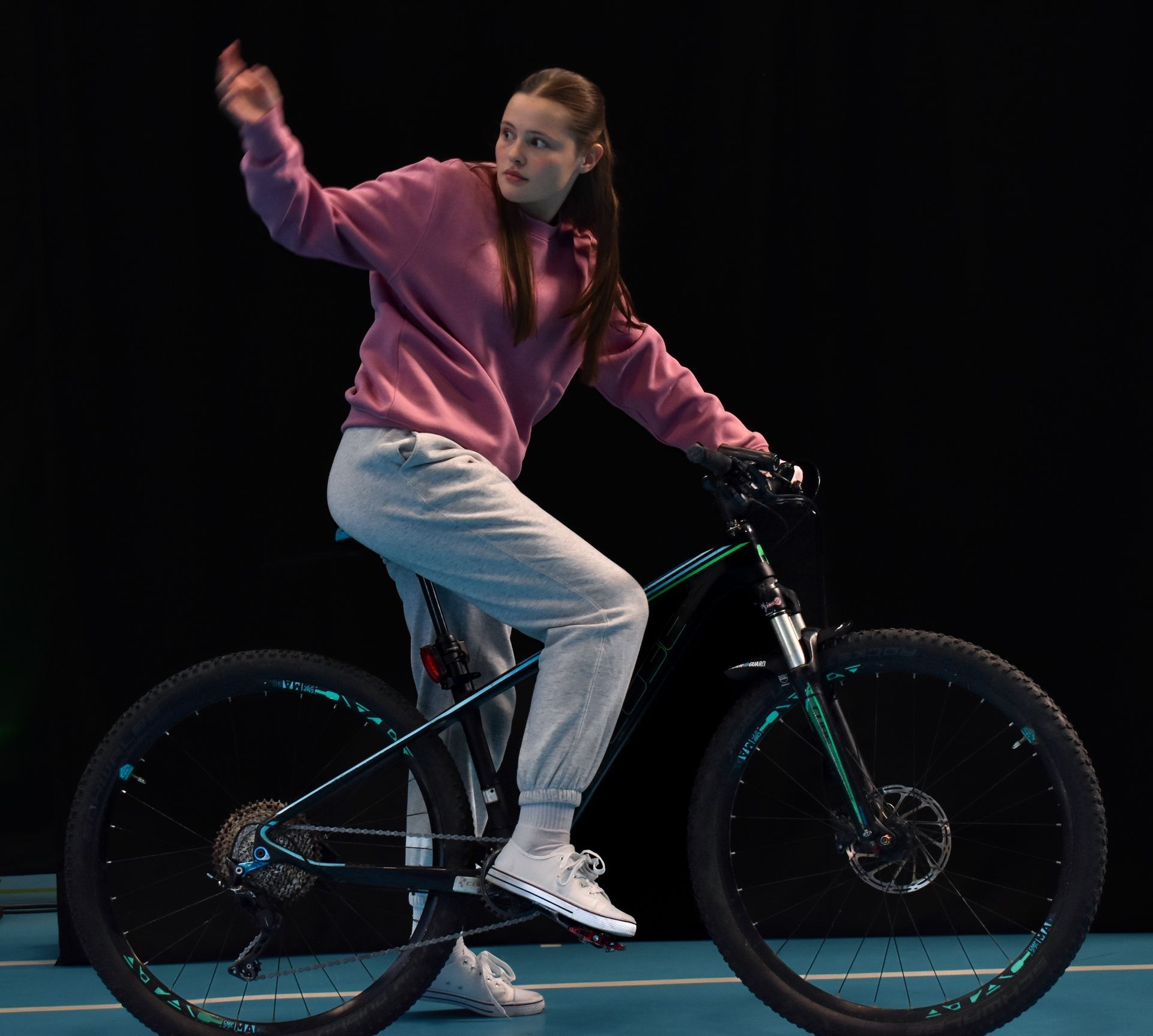 A teenage girl in a pink hoodie riding a bike against a dark studio backdrop
