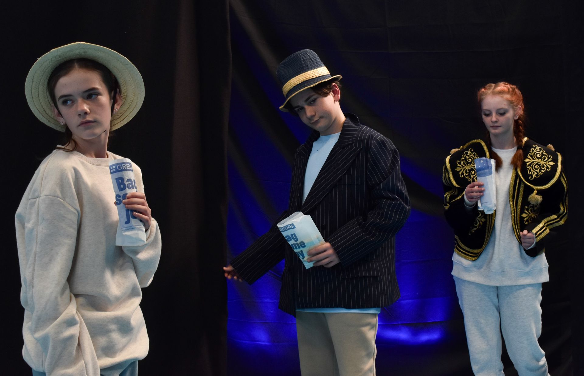 Three teenage performers in hats posing onstage under blue lights, holding props and wearing white and dark outfits