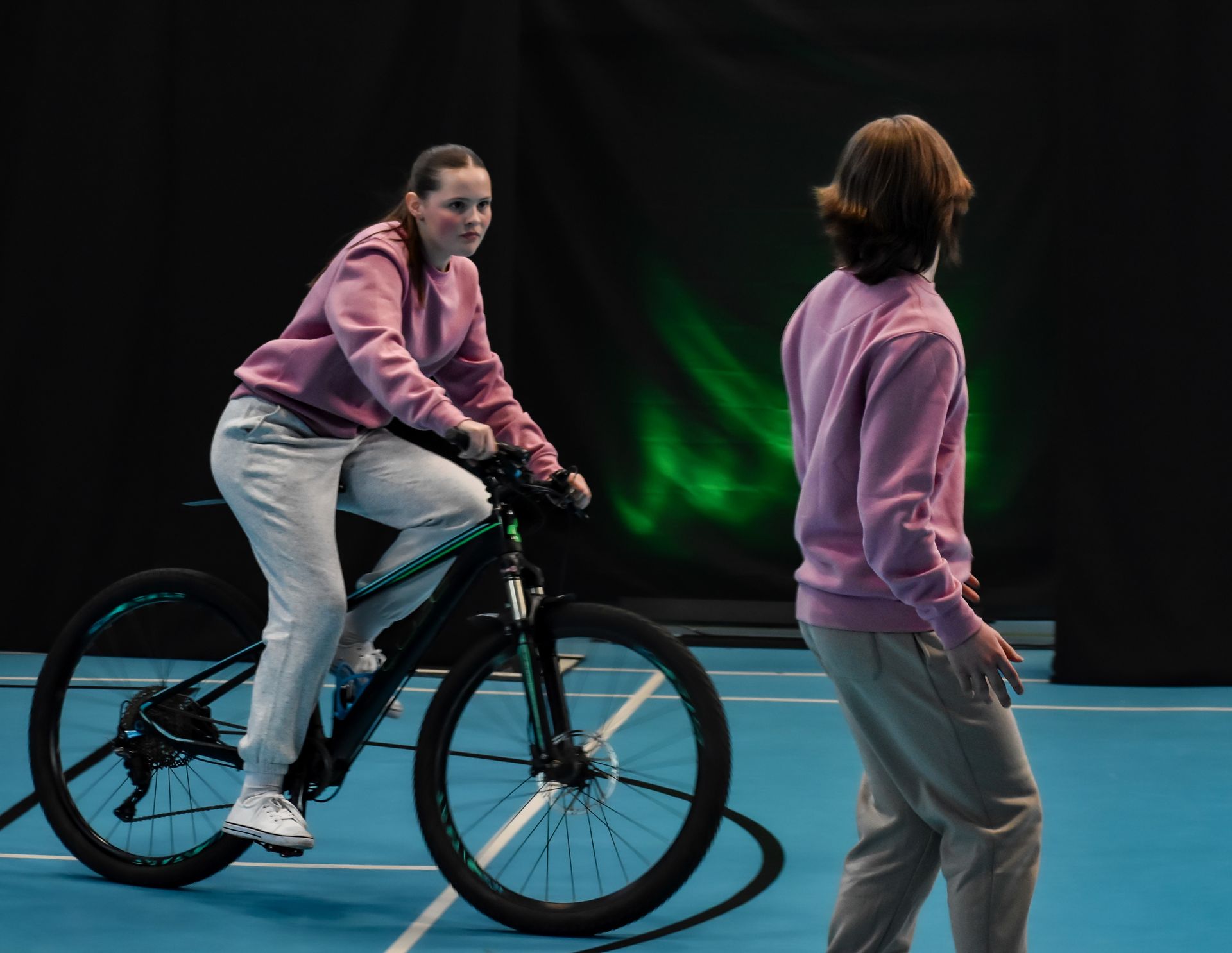 Two people in pink tops in a gym hall, one riding a bicycle and one standing nearby under dark lighting