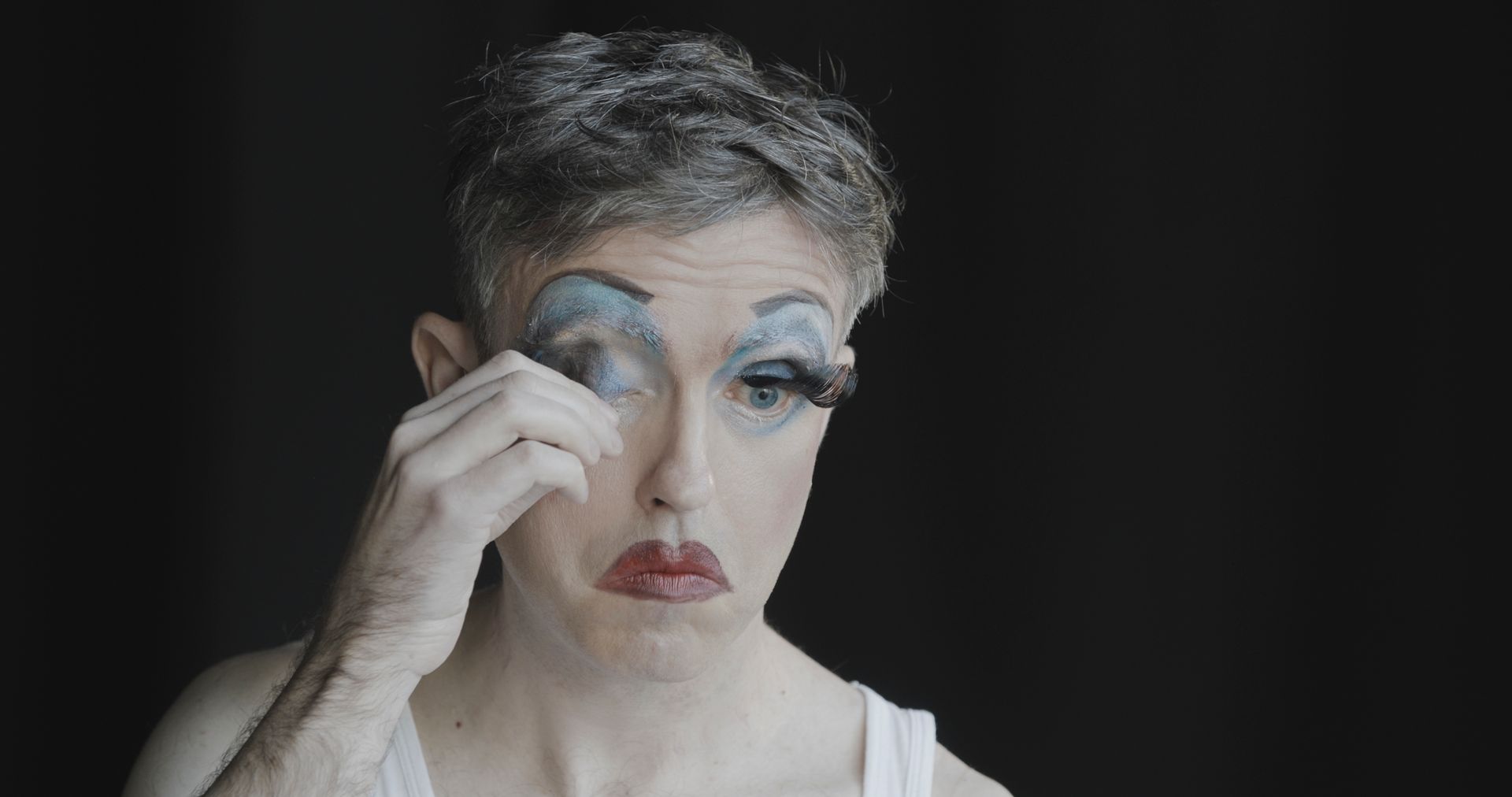 A man with makeup on his face is removing a false eyelash.