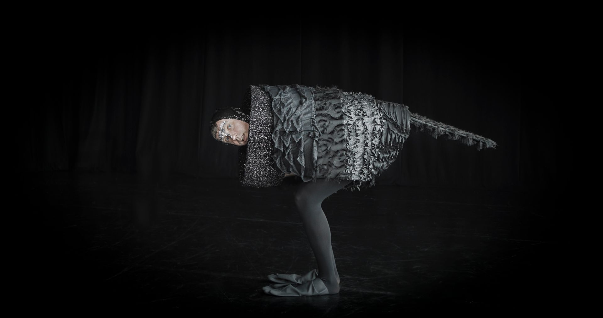 A man in a black crow costume is standing on one leg on a black background.