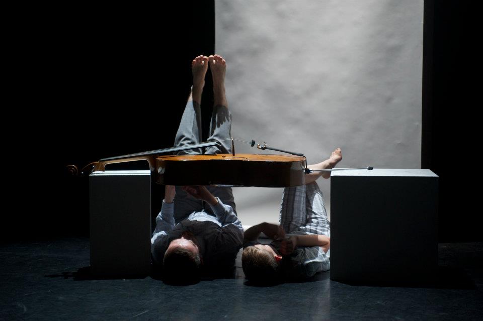 Two people are laying on the floor underneath a cello suspended between two blocks.