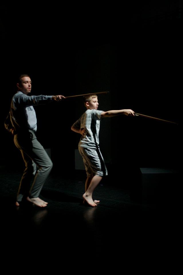 A man and a boy are pointing cello bows like sword in the dark.