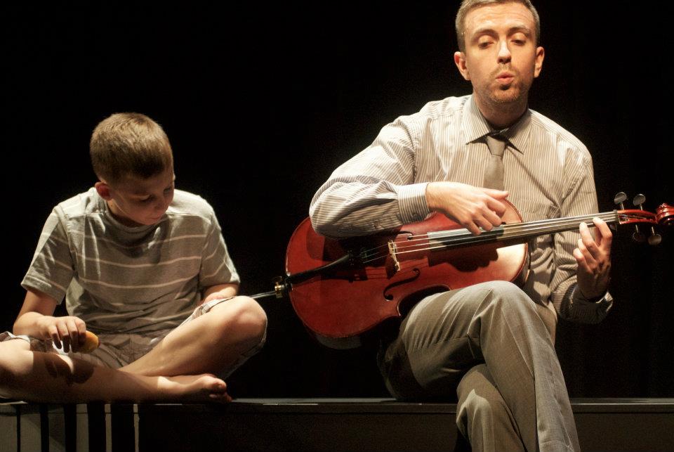 A man is playing a miniature cello while a boy sits next to him