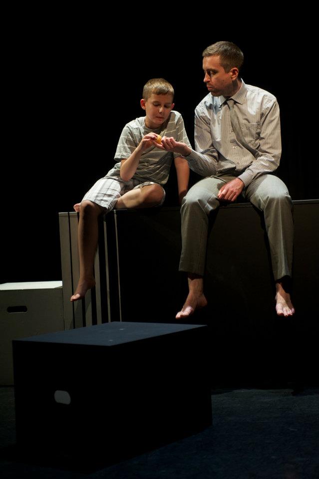 A man and a boy are sitting on a box on a stage sharing an apple.