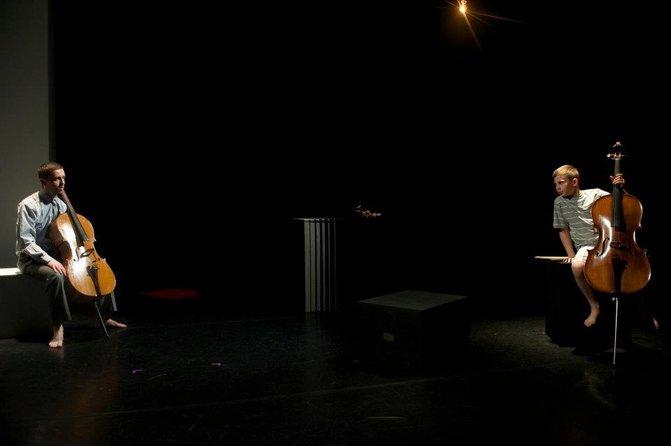 Two men are playing cello on a stage in a dark room.
