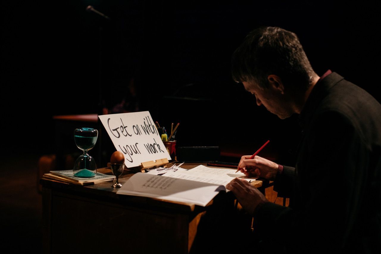 Man writing at a desk by lamp light, with papers and a timer in a dark room