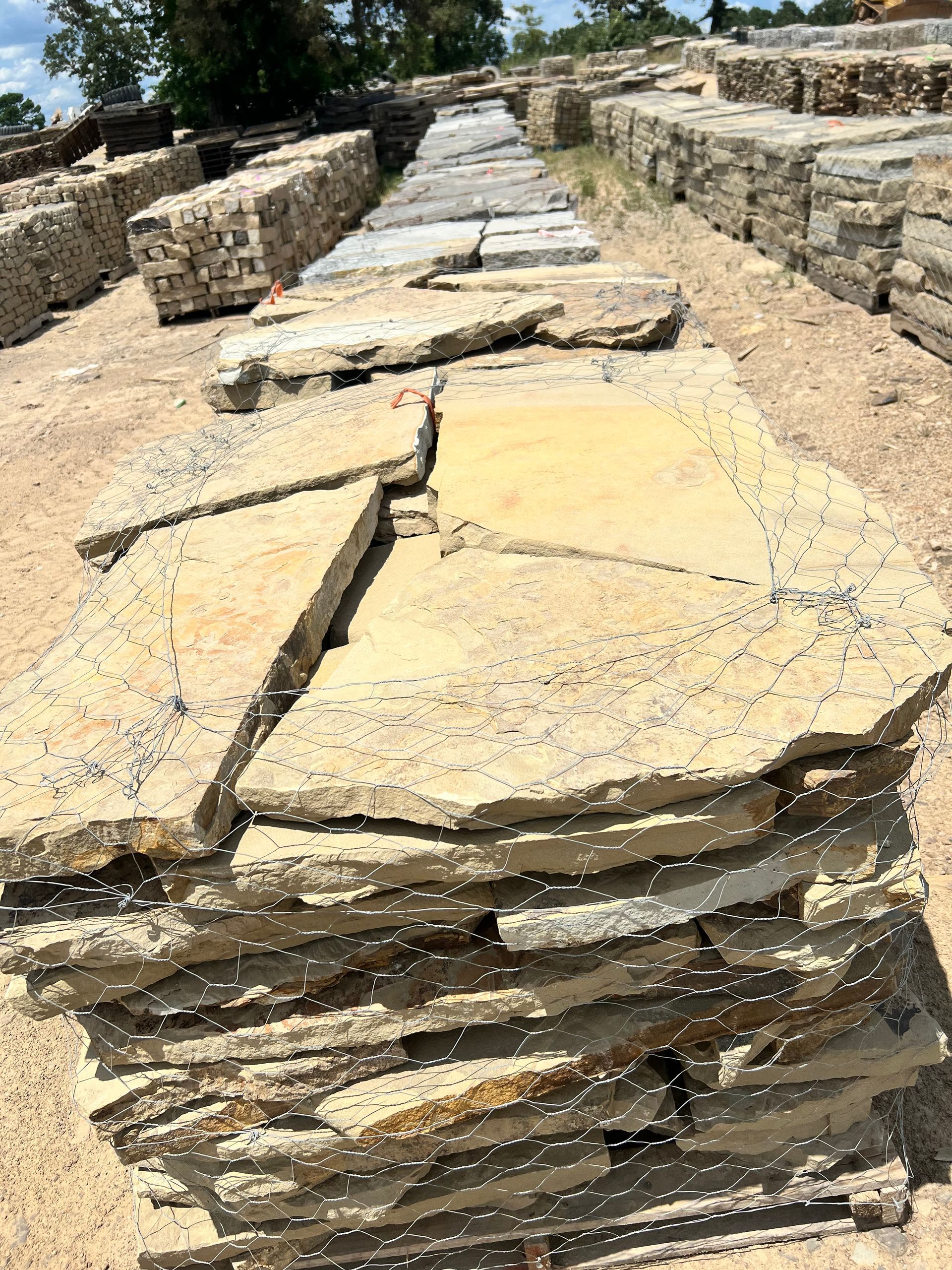 A stack of tan flagstone slabs sits in a stone yard, with rows of other masonry products extending into the background.