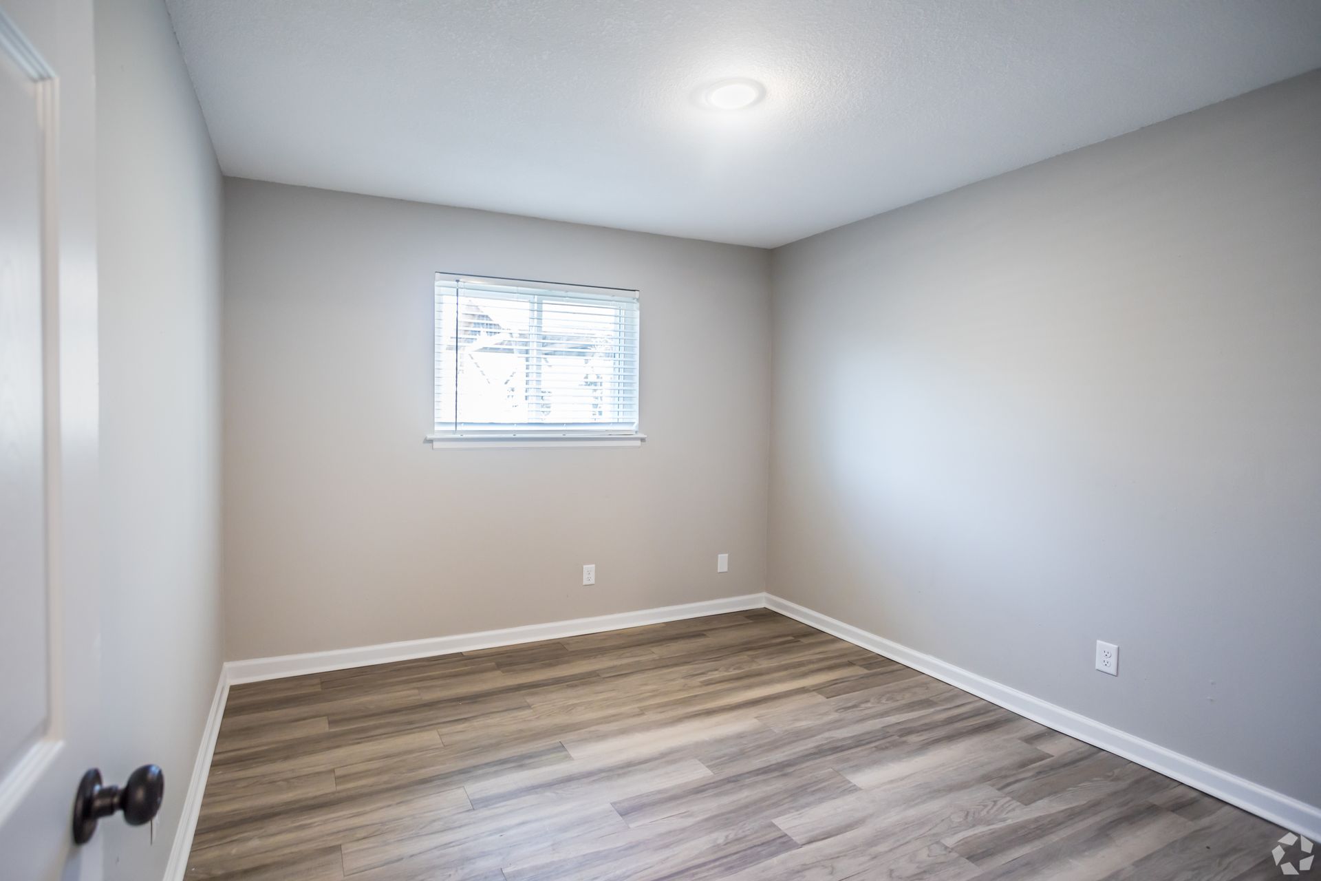 Empty room with grey walls, window, hardwood floors, and overhead light.