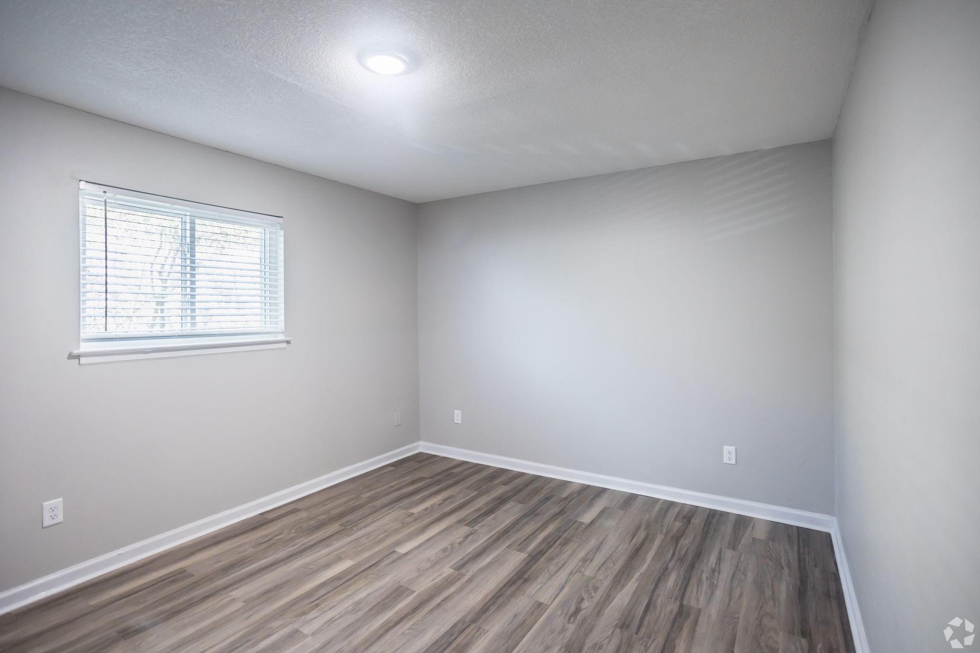 Empty bedroom with wood-look floor, window with blinds, and light gray walls.