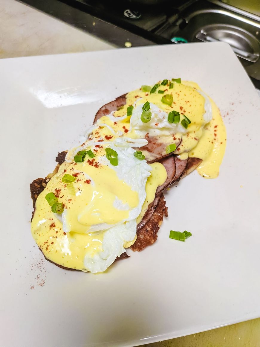 Eggs Benedict on a white plate, topped with yellow hollandaise sauce and green garnish.