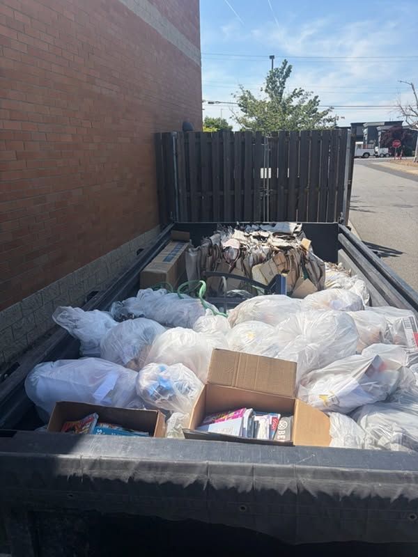 Dumpster overflowing with white trash bags and cardboard beside a brick wall and black fence outside.