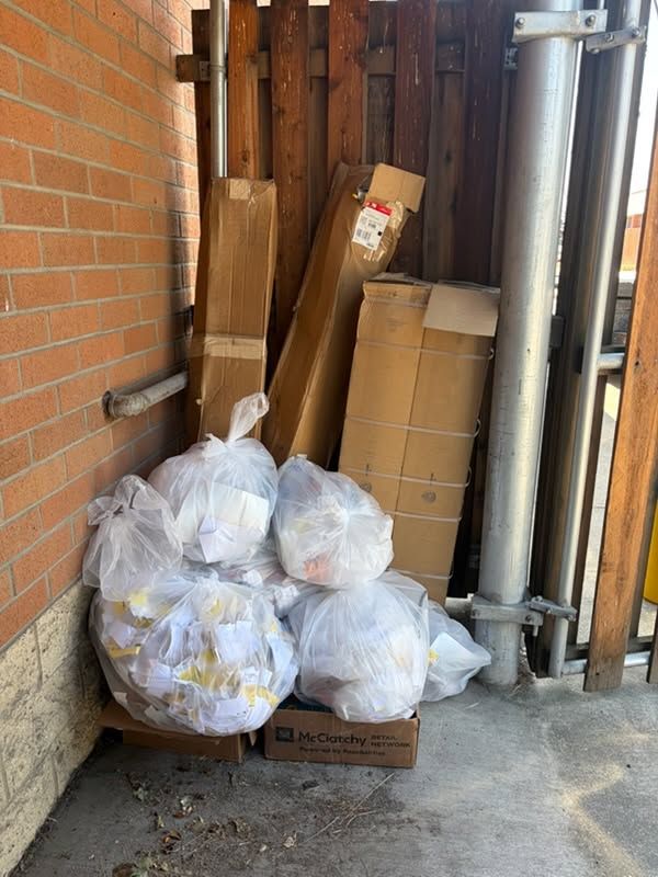 Trash bags and cardboard boxes stacked in a narrow alley beside a brick wall and metal pipes.