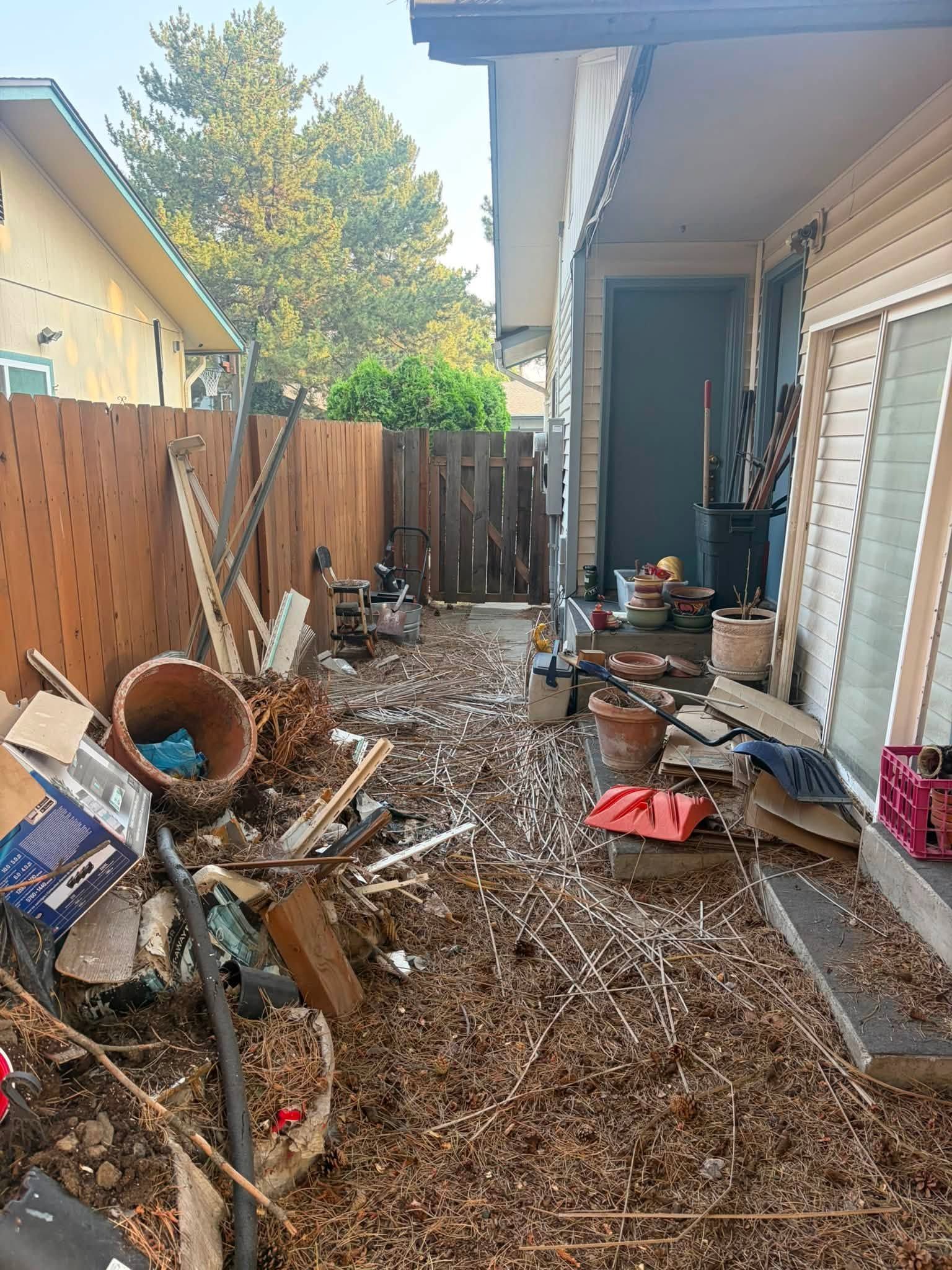 Narrow backyard cluttered with debris, wood scraps, and tools beside a house and wooden fence.