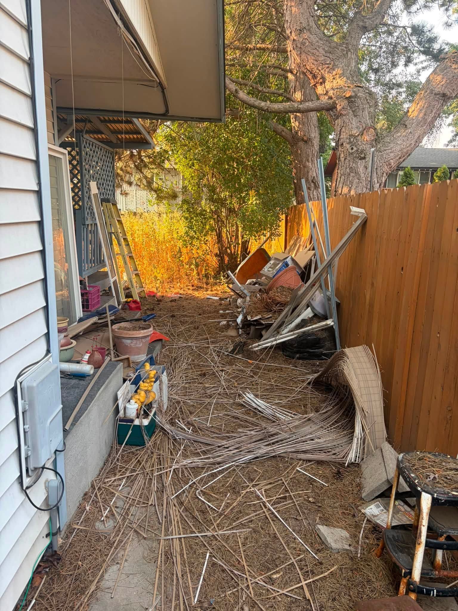 Narrow backyard side yard with debris, tools, and fallen branches beside a wooden fence and house wall.
