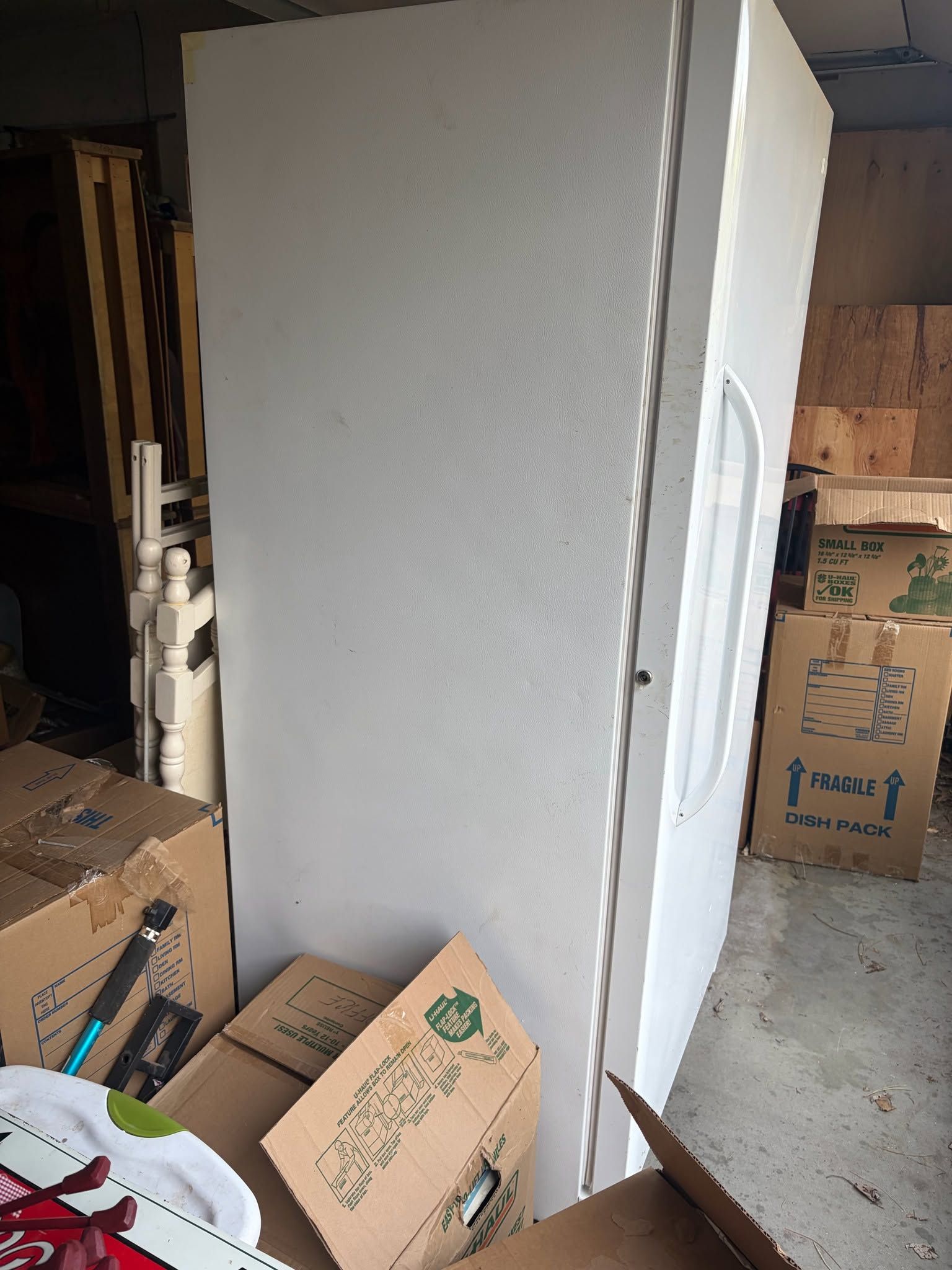 White refrigerator standing upright in a cluttered garage with moving boxes around it