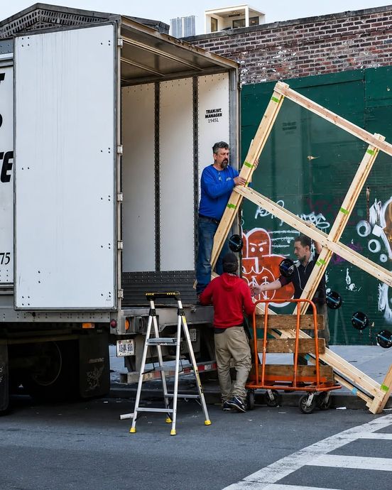 Two workers move a large wooden frame beside a truck and ladders in a narrow alley.