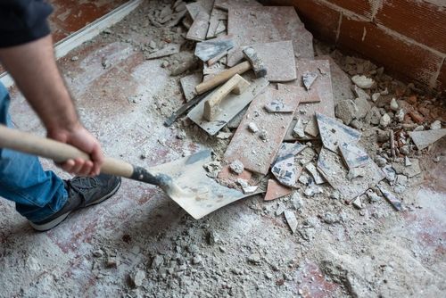 Person shoveling debris and broken tile in a dusty room under construction