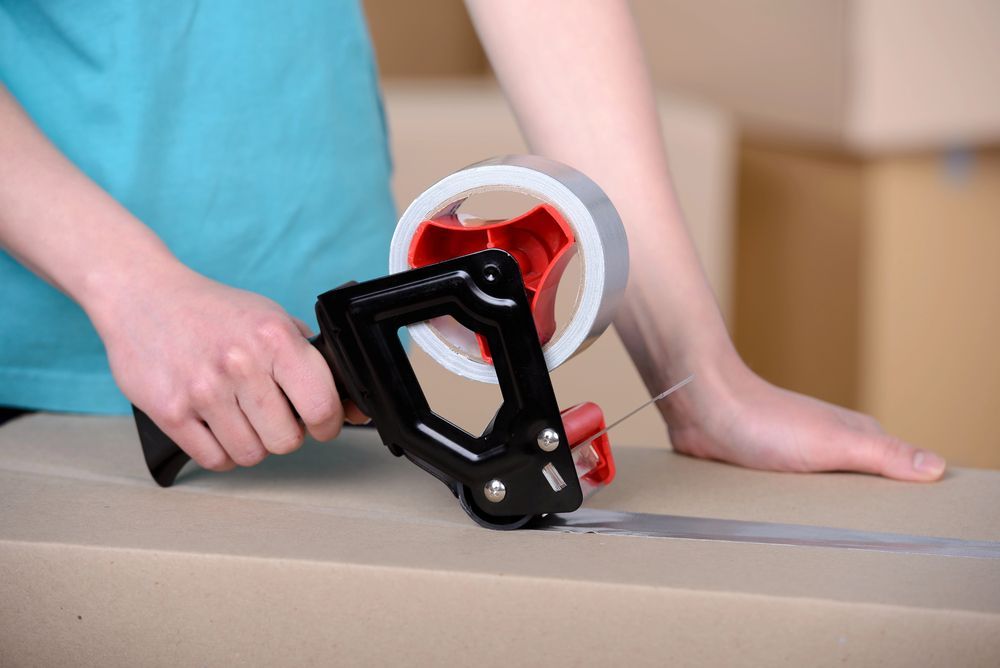 Persona sellando una caja de cartón con cinta de embalaje usando un dispensador de cinta manual.