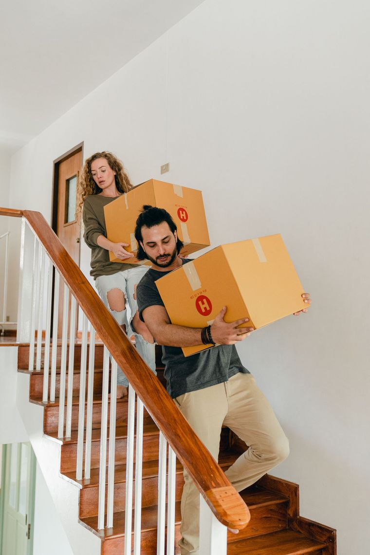 Dos personas bajan cajas de cartón de mudanza por una escalera en una casa luminosa.