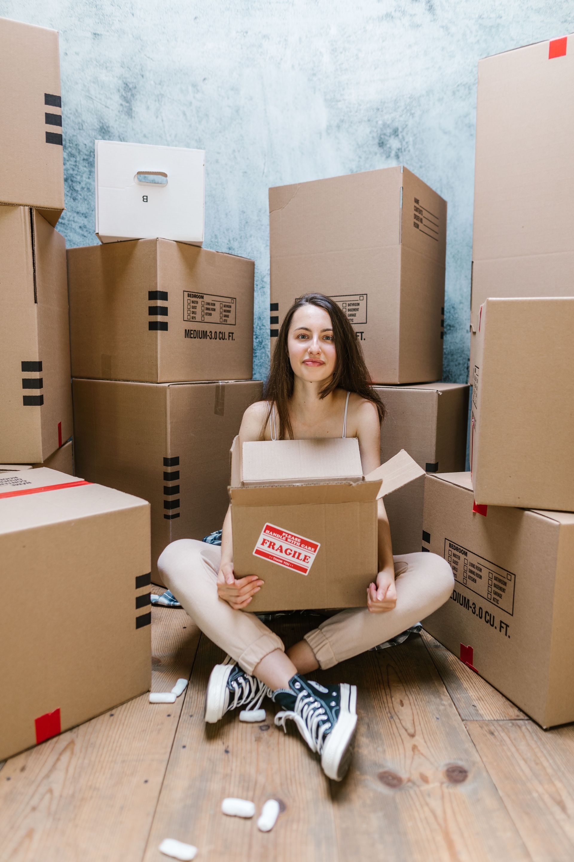 Una persona sentada entre cajas de cartón apiladas, sosteniendo una caja abierta y sonriendo en una habitación con suelo de madera.