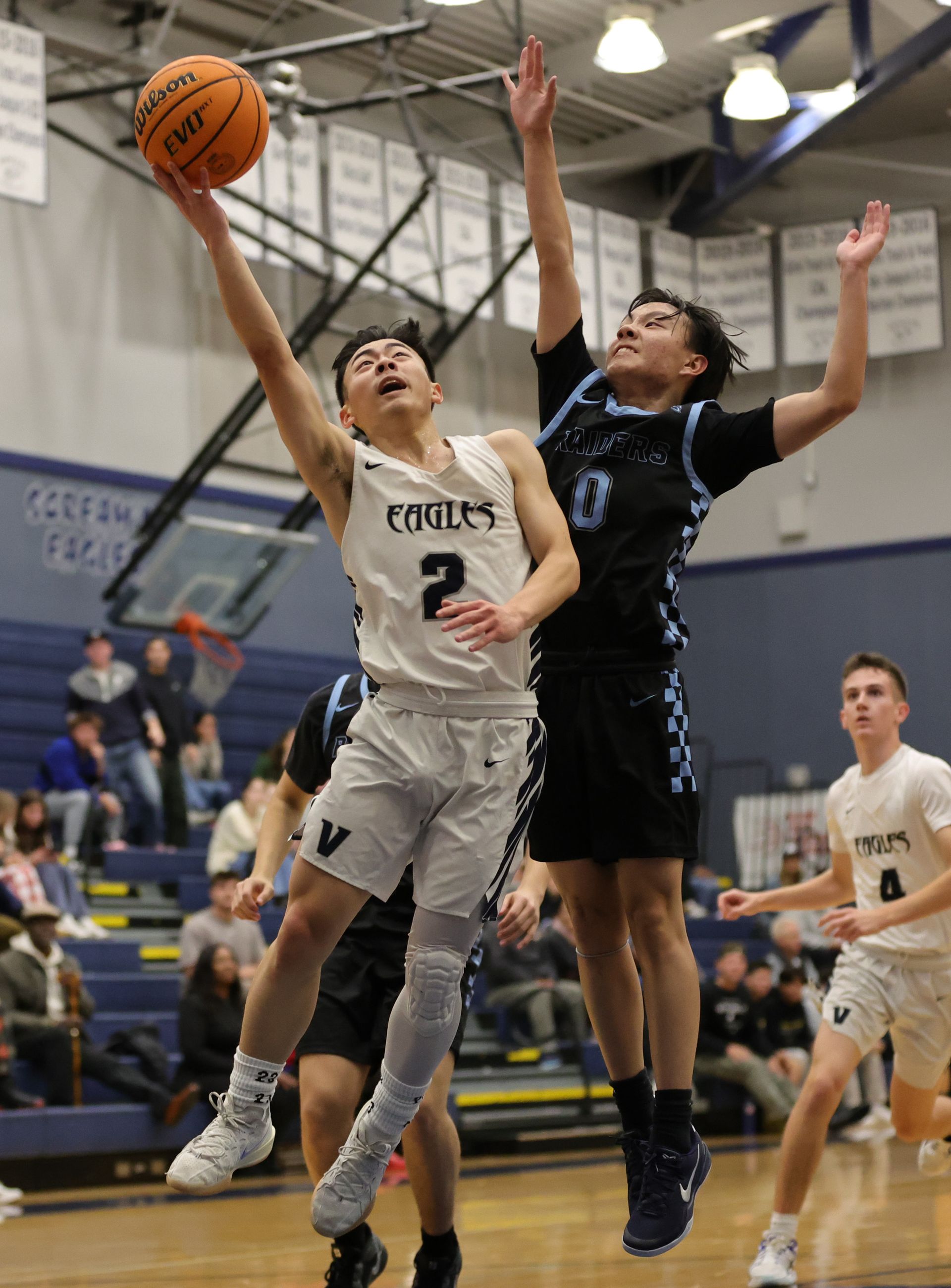 Basketball player in white jersey shoots while another player jumps to block the shot.