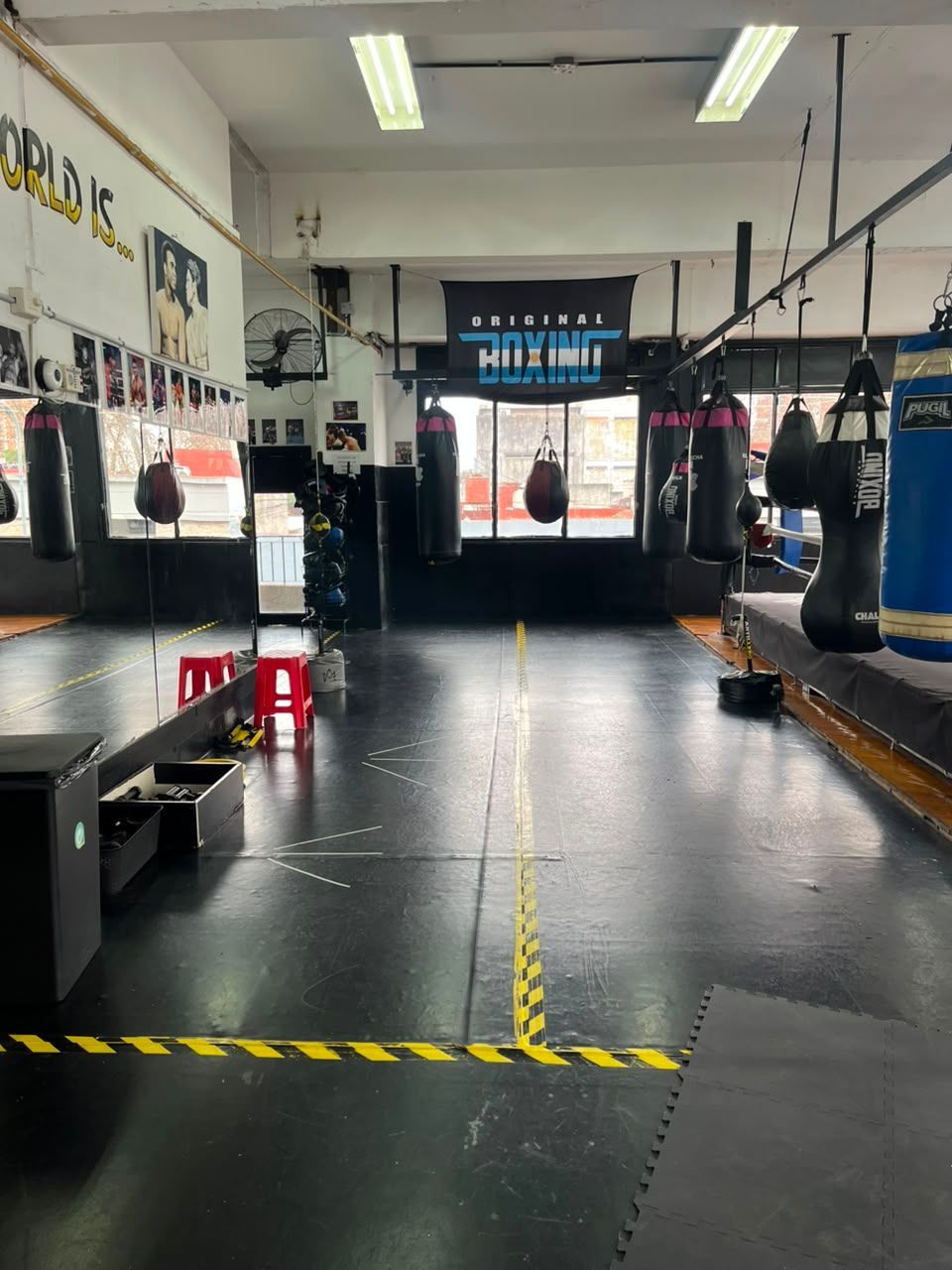Gimnasio de boxeo vacío con sacos de boxeo colgados, espejos y un suelo de estera negra.