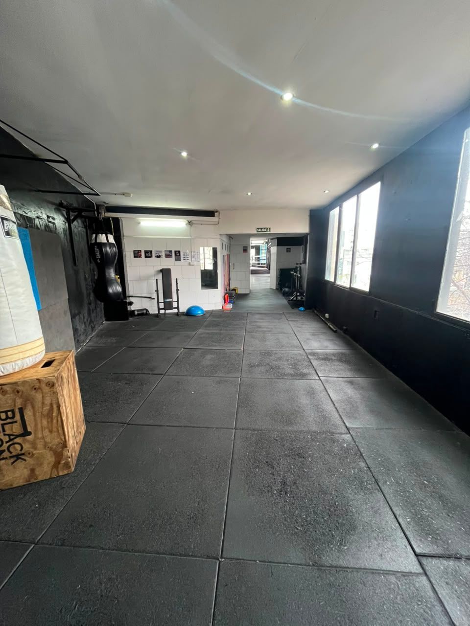 Gimnasio interior vacío con suelo de goma negra, paredes blancas y grandes ventanales a la derecha.