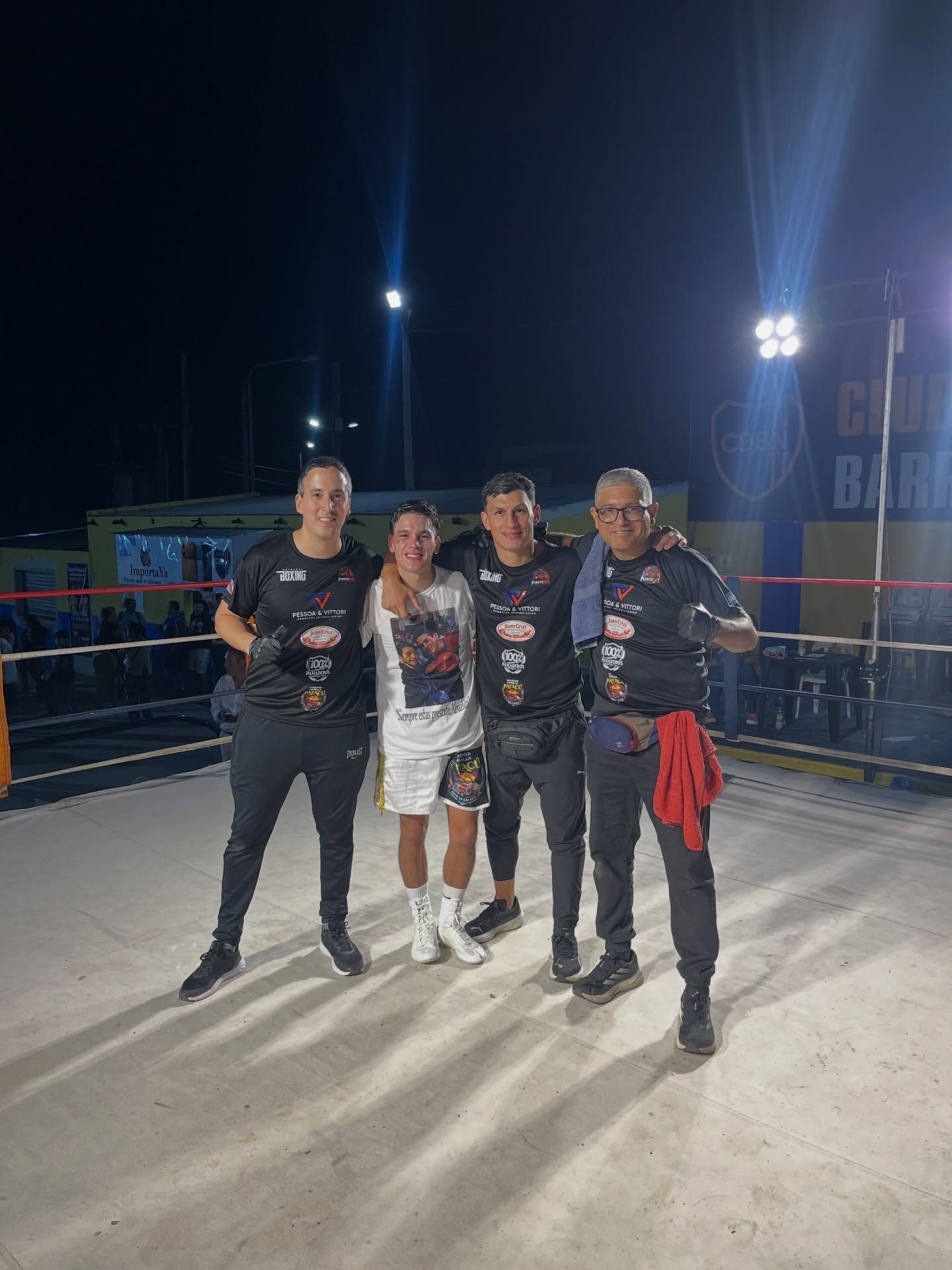 Cuatro jugadores de hockey posan de noche en una pista de hielo al aire libre, vestidos con camisetas y patines oscuros.
