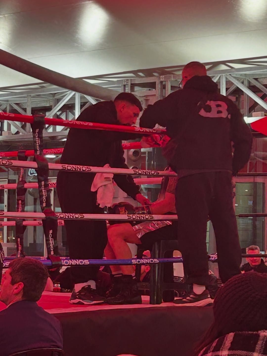 Rincón del ring de boxeo con un boxeador sentado, un entrenador de pie a su lado y cuerdas rojas bajo luces brillantes.