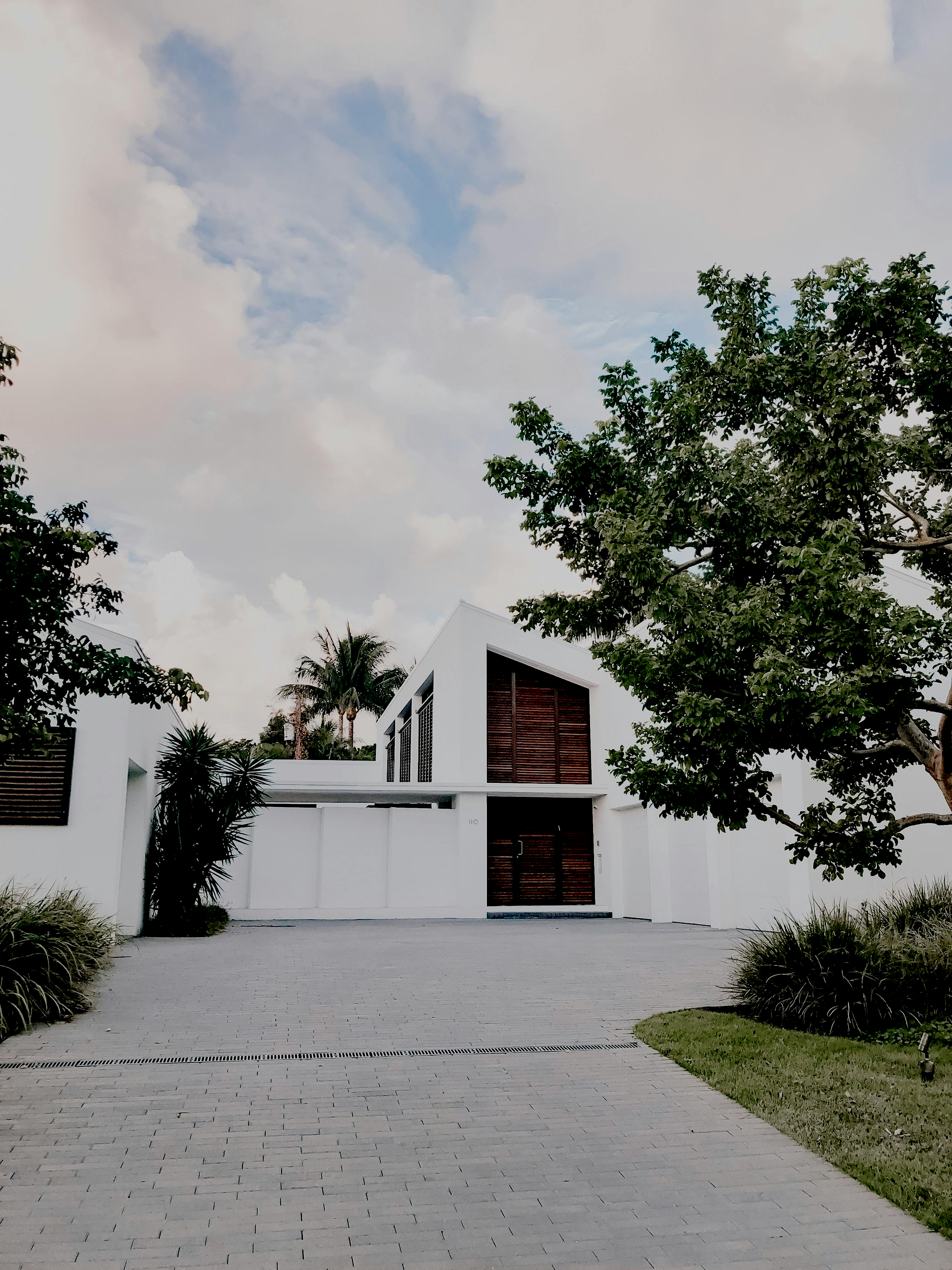 Maison blanche moderne avec allée en briques, encadrée d'arbres et de verdure sous un ciel nuageux
