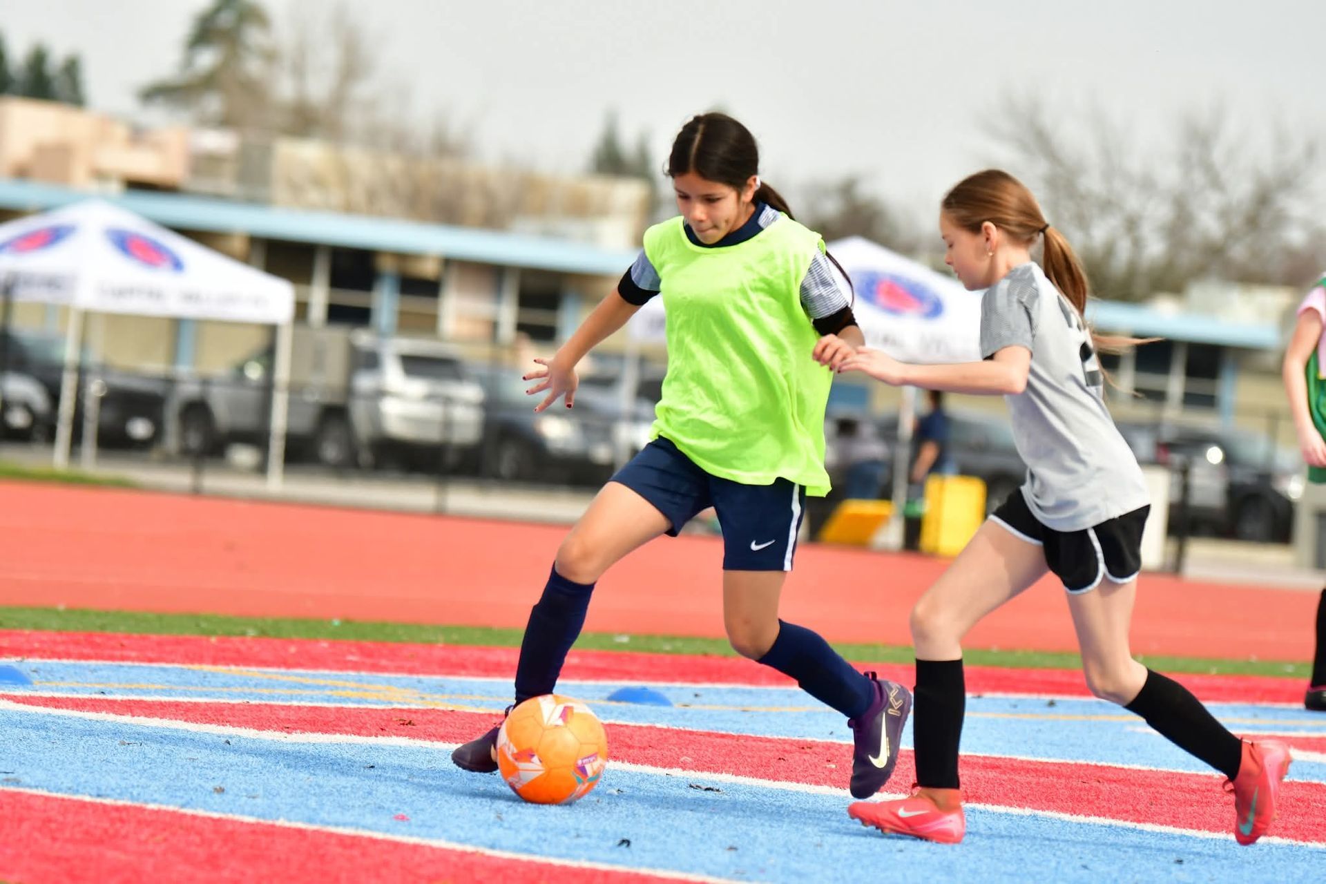 Two soccer players in athletic gear dribble and compete for control of a soccer ball on a red and blue surface outdoors.