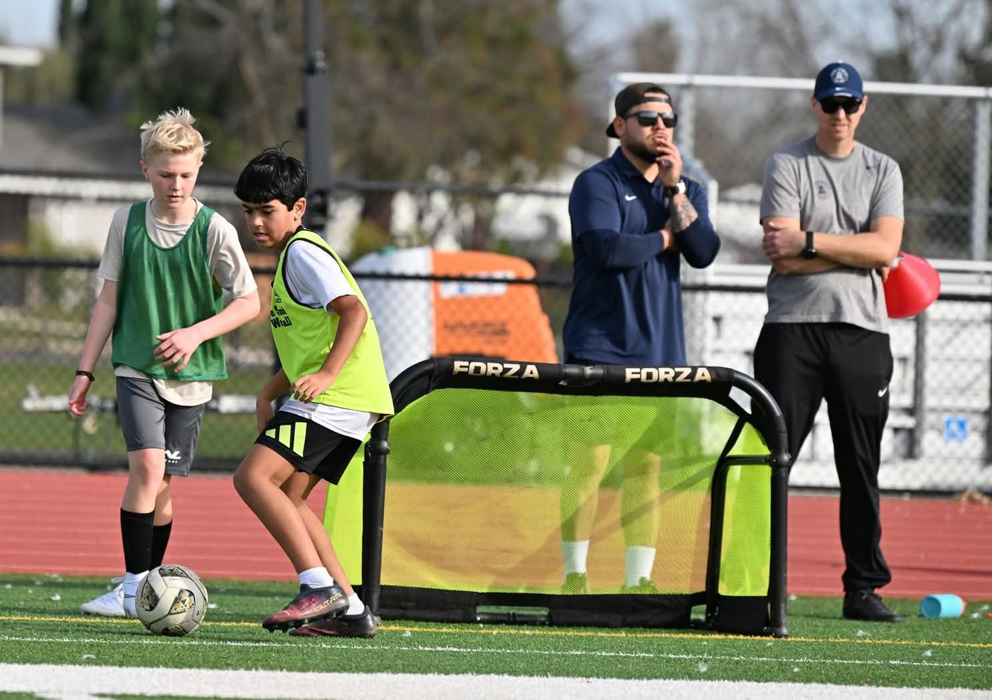 Two children play soccer near a small goal on a field while two coaches stand behind them watching.