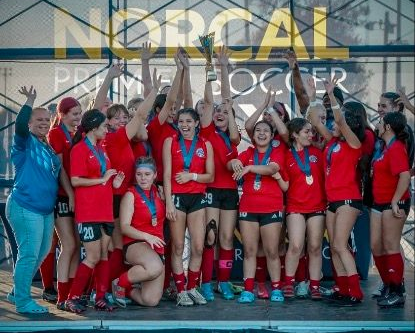 A soccer team in red uniforms cheering and holding a trophy under a NorCal Premier Soccer banner.