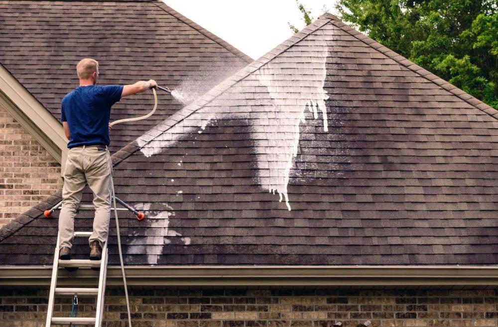 A man is standing on a ladder cleaning the roof of a house.
