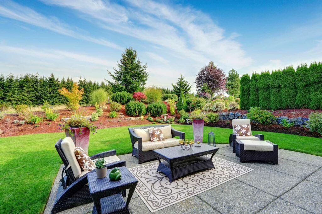 A patio with a couch , chairs , table and rug in a backyard.