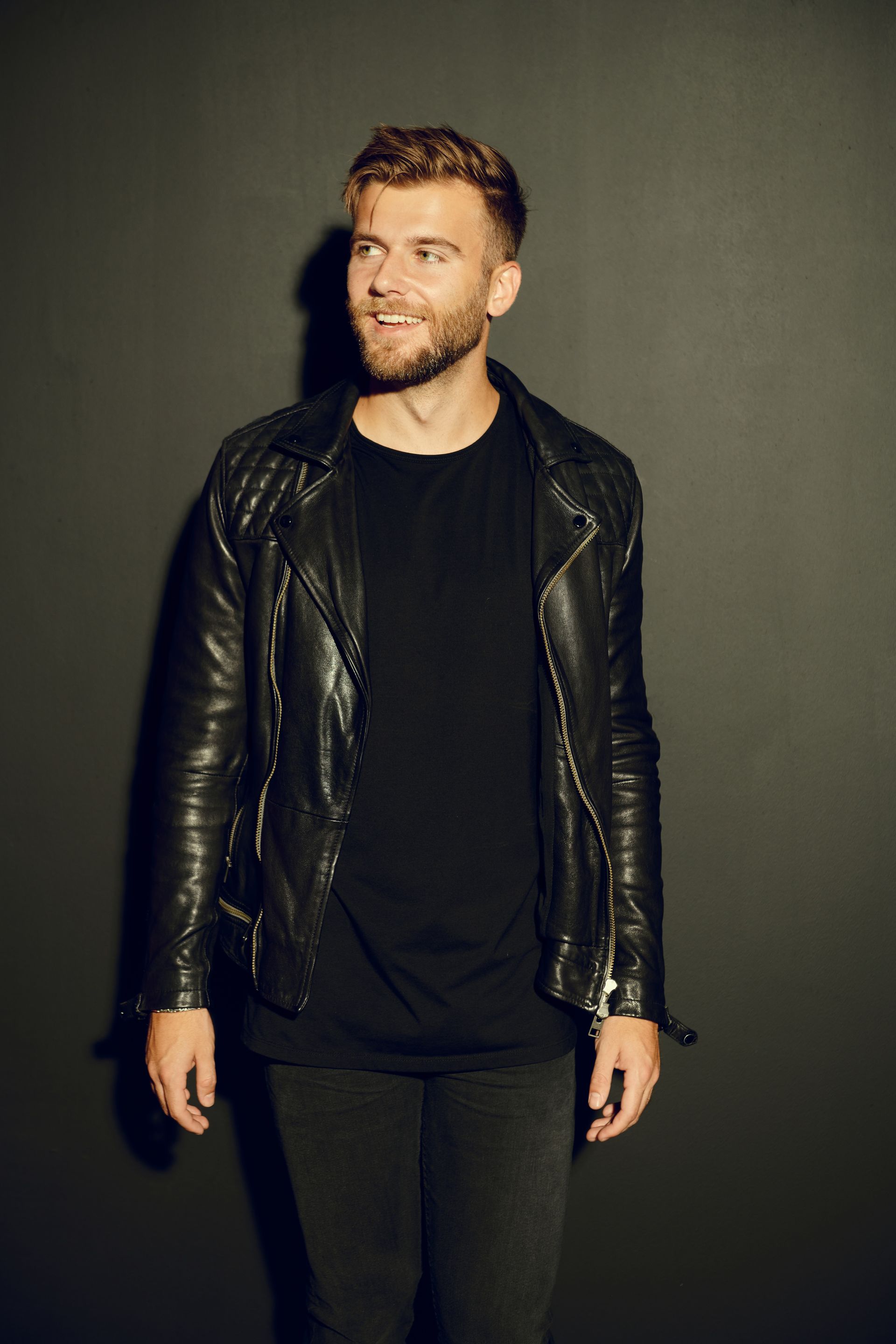 A man wearing a black leather jacket and a black shirt is standing in front of a black wall.