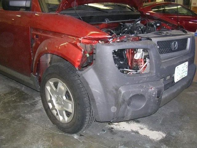 Red SUV with severe front-end damage in a garage, hood open and front bumper broken