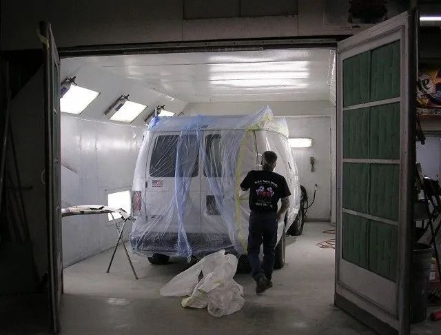 Person in a workshop sprays a white vehicle under plastic sheeting inside a garage