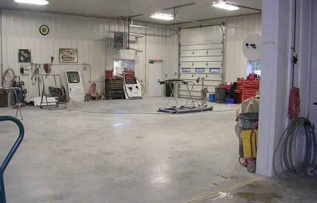Empty industrial garage with concrete floor, shelves, tools, and equipment under fluorescent lights