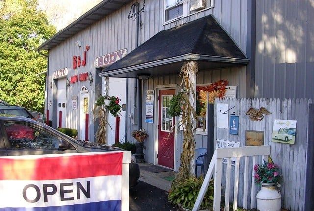 Small shop exterior with an OPEN sign, gray siding, red trim, and potted flowers by the entrance.