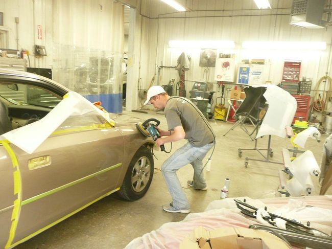 Two workers sand and prep a brown car in an auto body shop with covered vehicles nearby