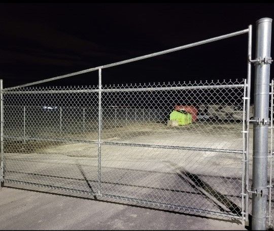 Un portail en grillage bloque un terrain vague la nuit, avec une tente lumineuse au néon en arrière-plan.
