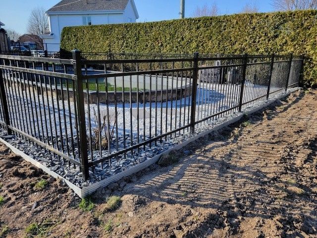 Clôture en métal noir entourant une petite cour avec une terrasse circulaire en béton, bordée par un chemin de terre et une haute haie.
