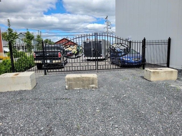 Parking en gravier avec un portail noir, des barrières en béton et une voiture bleue garée à côté d'un bâtiment.