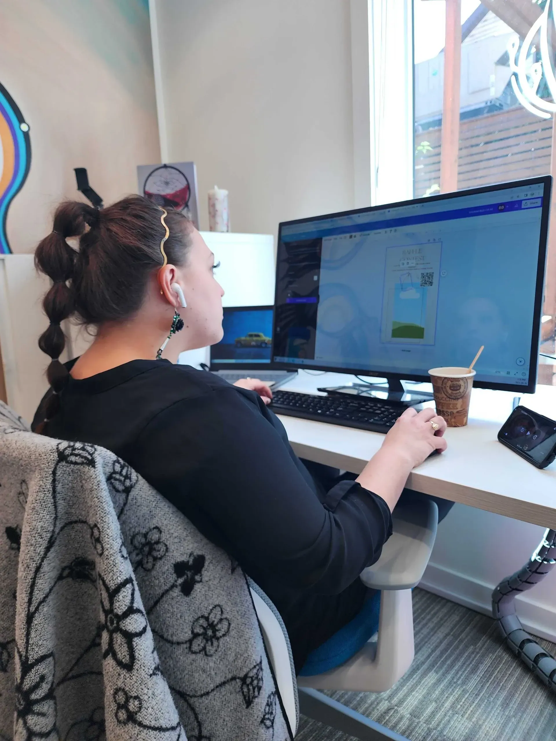 Woman at computer, right hand on mouse.  Wearing headphones, seated at a desk near a window. Coffee cup and phone present.