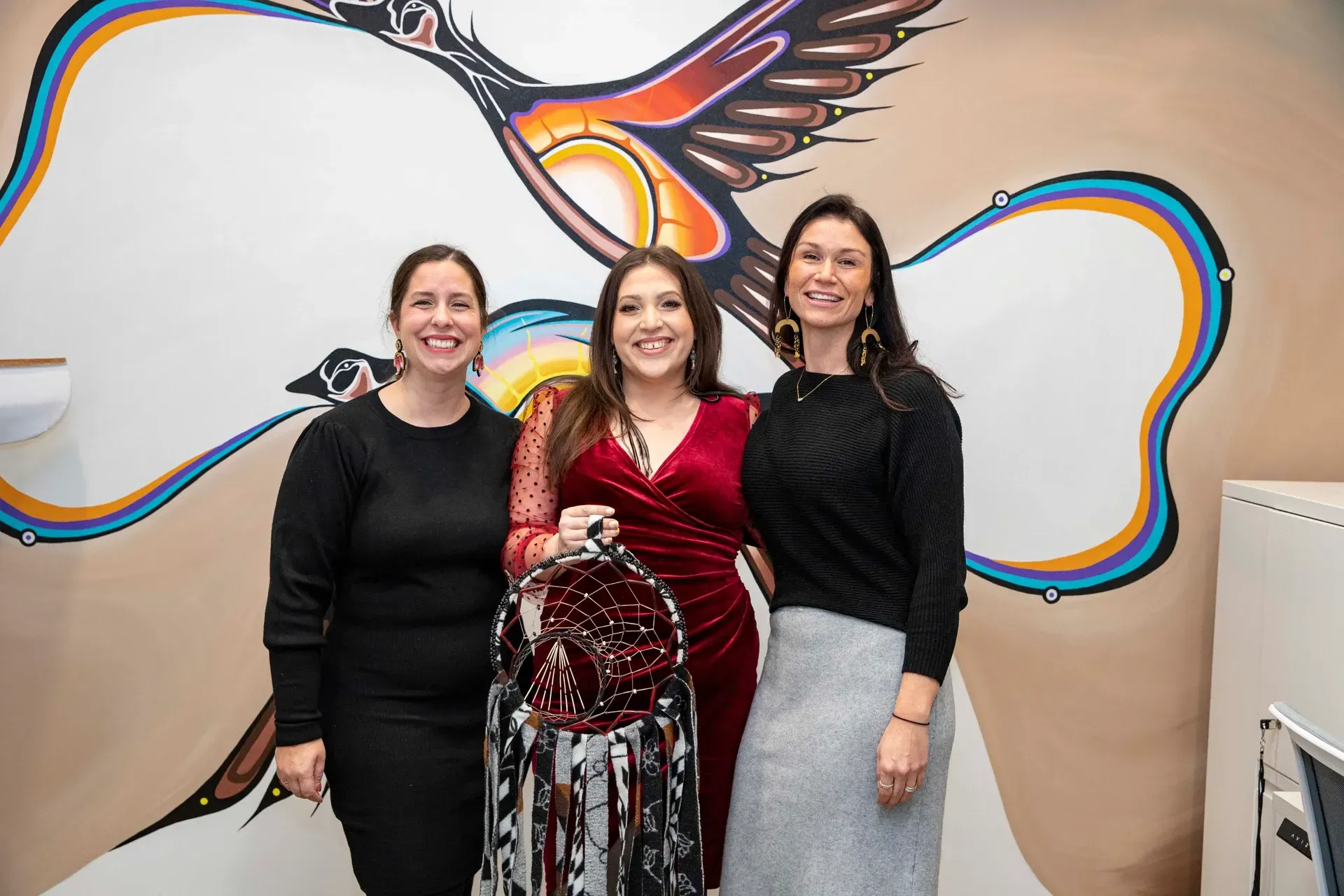 Three women posing with a decorated item in front of a mural with bird imagery.